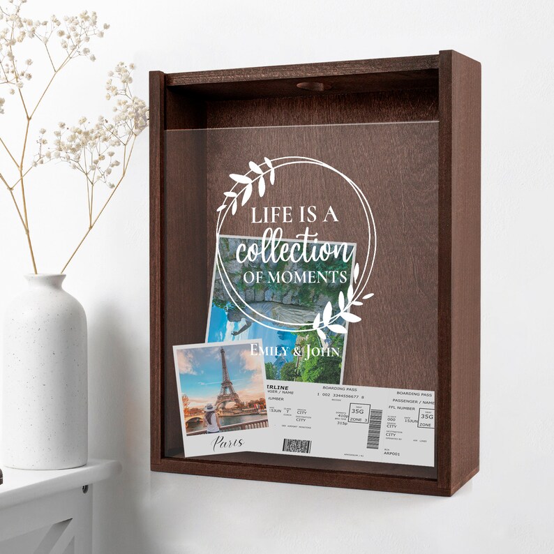 Peut inclure: Une bo&icirc;te &agrave; ombre en bois brun avec un devant en verre transparent. La bo&icirc;te contient deux photos et une carte d'embarquement. Les photos sont de la tour Eiffel &agrave; Paris. La carte d'embarquement est pour un vol vers Paris. La bo&icirc;te a un autocollant en vinyle blanc avec le texte "Life is a collection of moments" et un motif floral.