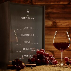 Puede incluir: Un estante de vino de madera con un panel de vidrio que muestra una escala de vino con los niveles: Kristin, Sommelier, Experto, Regular, Principiante. Un vaso de vino tinto y uvas est&aacute;n en primer plano.