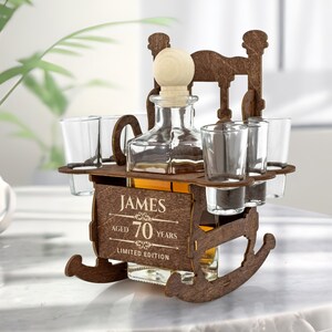 May include: Wooden rocking chair liquor dispenser with four shot glasses and a bottle of liquor. The dispenser is engraved with the text "James Aged 70 Years Limited Edition".