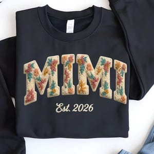 May include: A black sweatshirt with the word "MIMI" in a floral design. The text "Est. 2026" is below the word "MIMI". The sweatshirt is displayed with a pair of blue jeans and a black and white striped scarf.