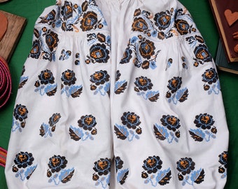 May include: White embroidered blouse with a floral pattern in black, blue, and orange. The blouse has long sleeves and a gathered neckline.