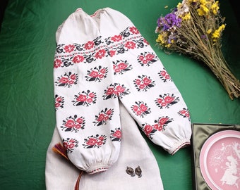 May include: A white embroidered blouse with red and black floral patterns, a white linen skirt with a brown belt, and two small decorative buttons.