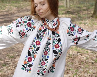 May include: White embroidered tunic with colorful floral patterns. The long-sleeved garment features intricate embroidery in red, blue, yellow, and green. A brown beaded necklace is worn with the tunic. The background is a blurred outdoor scene.