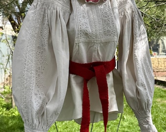 May include: An off-white long-sleeve blouse featuring intricate white eyelet embroidery, a ruffled collar with red stitching, and voluminous sleeves with ruffled cuffs. A bright red woven sash is tied around the waist of the garment.