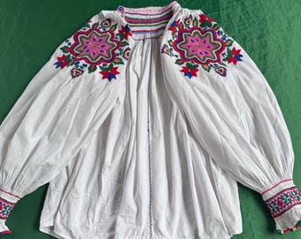 May include: A white long-sleeved blouse featuring vibrant multi-colored floral embroidery on the shoulders and cuffs. The garment has a gathered neckline with a colorful stitched trim and full, billowy sleeves that taper into ruffled, embroidered wrist cuffs.