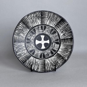 May include: A black and white decorative plate with a cross design in the center. The plate features a radial pattern with alternating black and white lines. The plate is displayed on a black stand against a gray background.