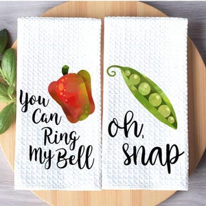 May include: Two white kitchen towels with a waffle weave texture. One towel features a red and yellow bell pepper with the text "You Can Ring my Bell". The other towel has a green pea pod with the text "Oh, snap".