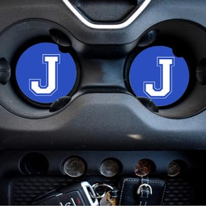 May include: Two blue car coasters with a white letter J on each. The coasters are in the cup holders of a car.