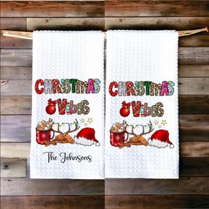 May include: Two white kitchen towels with a Christmas theme. The towels feature the text "Christmas Vibes" in colorful patchwork letters, a red mug with marshmallows and a candy cane, two gingerbread cookies, and a Santa hat. The towel on the left has the text "The Johnsons" printed below the design.
