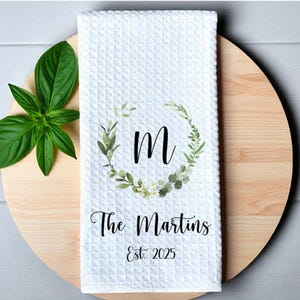May include: White waffle-weave kitchen towel with a botanical wreath around the letter "M". The text "The Martins Est. 2025" is printed below the letter. A sprig of green leaves is on the left side.