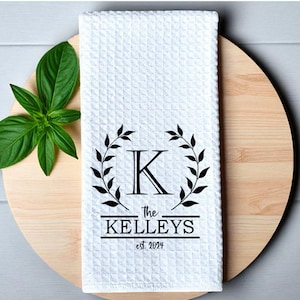 May include: White waffle-weave kitchen towel with a black monogram "K" inside a laurel wreath design. The text "The Kelleys est 2024" is printed below the wreath. The towel is displayed on a wooden cutting board with green leaves.