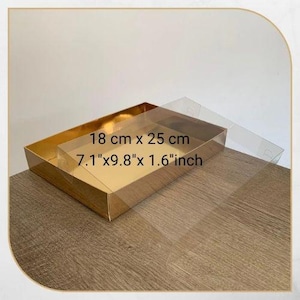 Puede incluir: Una caja rectangular dorada con tapa transparente, mide 18 cm x 25 cm x 4 cm. La caja está abierta, revelando un interior dorado. La tapa transparente cubre parcialmente la caja. La caja está sobre una superficie de madera.