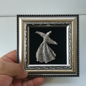 May include: A silver framed picture with a black velvet background featuring a silver 3D sculpture of a whirling dervish.