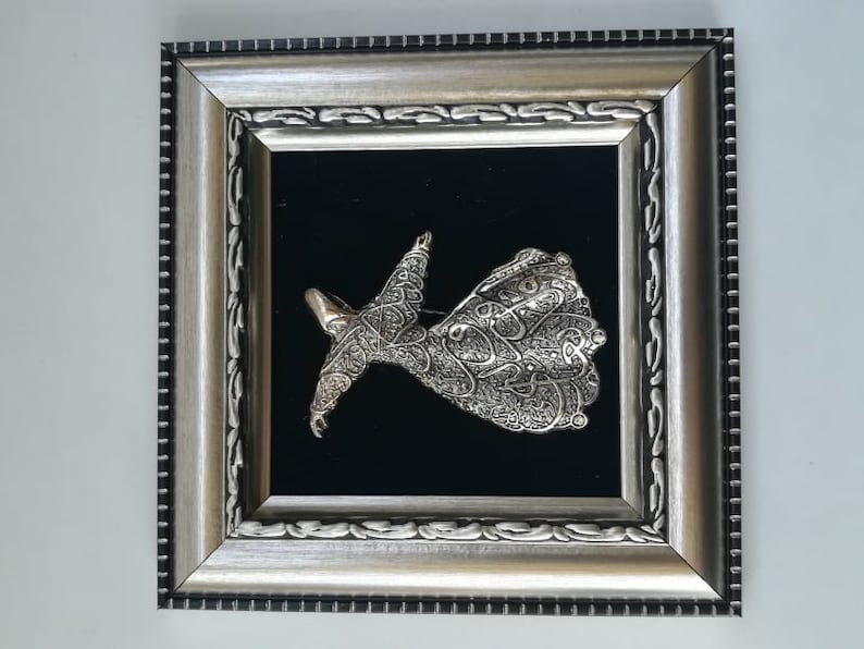 May include: A silver-colored, framed wall art piece depicting a whirling dervish figure with intricate calligraphy details. The figure is set against a black velvet background.