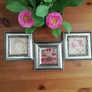 May include: Three silver picture frames with ornate designs, each containing a different embroidered piece of fabric. The frames are arranged in a row on a wooden surface.