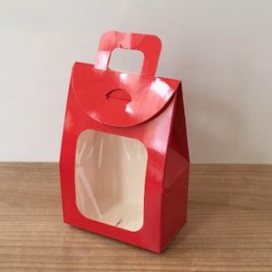 Puede incluir: Una caja de regalo roja con una ventana y un asa. La caja está hecha de cartón y tiene un acabado brillante.