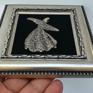 May include: A silver-colored metal sculpture of a whirling dervish, framed in a silver-colored frame with black velvet backing.