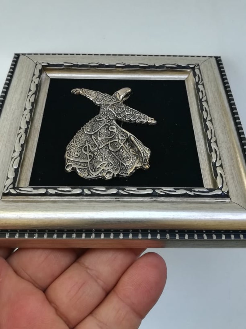 May include: A silver-colored metal sculpture of a whirling dervish, framed in a silver-colored frame with black velvet backing.