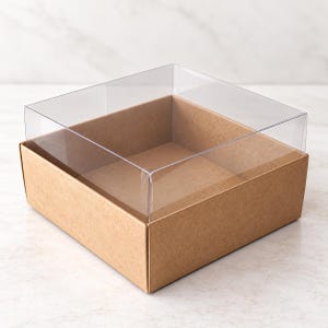 Könnte beinhalten: Eine quadratische Schachtel mit einem braunen Kartonboden mit vier Seiten und einem durchsichtigen Kunststoffdeckel, der oben aufliegt. Die Schachtel ist leer und hat ein schlichtes, funktionales Design mit klaren Linien und einem sichtbaren Innenraum.
