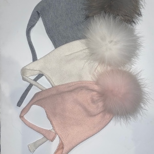 May include: Three knitted hats with fur pom-poms. The hats are gray, white, and pink. The pom-poms are gray, white, and pink.