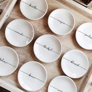 Könnte beinhalten: Ein Holztablett mit neun kleinen, weißen Keramikschalen. Jede Schale trägt eine schwarze Inschrift: "breathe", "inspire", "fearless", "namaste", "thankful", "blessed", "believe", "brave" und "warrior". Das Tablett ist von Willow Lane Collective.