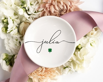 May include: White ceramic ring dish with the name "julia" written in black cursive script and a green four-leaf clover in the centre.