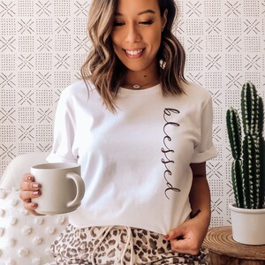 May include: White t-shirt with the word "blessed" in a vertical script. The person is wearing leopard print pants and holding a mug. A cactus in a white pot sits on a wooden table. The background has a white and black geometric pattern.