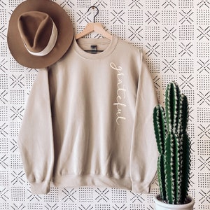 May include: A tan sweatshirt with the word "grateful" in cursive script. A brown hat with a white band is to the left. A cactus in a white pot sits on a wooden surface to the right. The background is a white and black patterned wall.