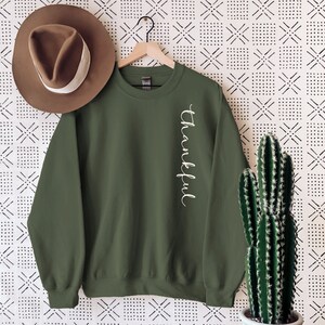 May include: An olive green sweatshirt with the word "Thankful" in white script down the side. A brown hat hangs to the left, and a cactus in a white pot sits on a wooden table. The background has a black and white geometric pattern.
