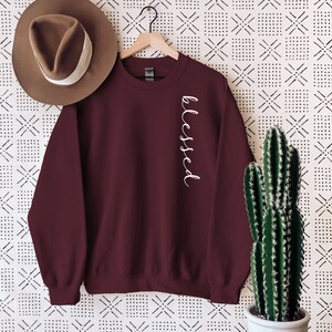 May include: A maroon sweatshirt with the word "blessed" in white script down the side. A brown hat with a white band hangs on the left. A cactus in a white pot sits on a wooden table to the right.