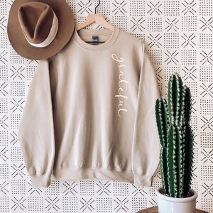 May include: A beige sweatshirt with the word "grateful" in cursive script on the side. A brown hat with a white band sits to the left of the sweatshirt. A cactus in a white pot is on the right.