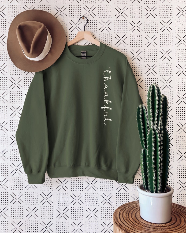 May include: An olive green sweatshirt with the word "Thankful" in white script down the side. A brown hat hangs to the left, and a cactus in a white pot sits on a wooden table. The background has a black and white geometric pattern.