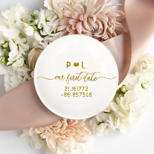 May include: A white ceramic dish with gold lettering and a heart symbol. The text reads "P ❤️ L our first date" with numbers below. The dish is surrounded by white and pink flowers and a pink ribbon.