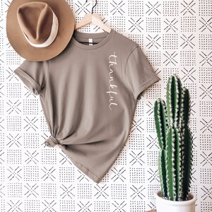 May include: A taupe t-shirt with the word "Thankful" in white script, hanging on a wooden hanger. A brown fedora hat rests on the shirt. A potted cactus sits on a wooden side table against a patterned wall.
