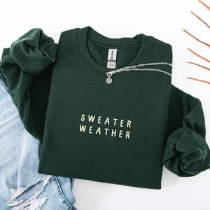 May include: A dark green sweatshirt with the words "SWEATER WEATHER" printed in white. A silver necklace with a pendant is draped across the sweatshirt. The sweatshirt is folded on top of a pair of blue jeans.