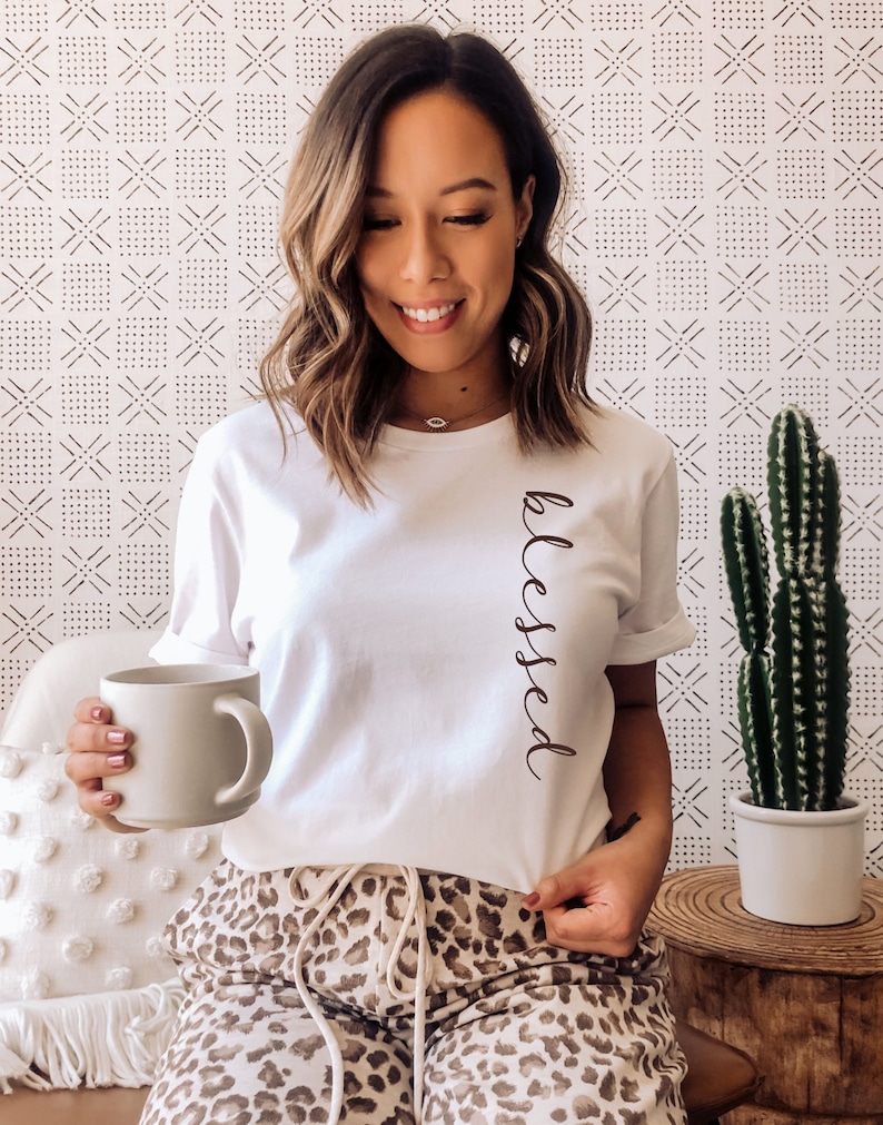 May include: White t-shirt with the word "blessed" in a vertical script. The person is wearing leopard print pants and holding a mug. A cactus in a white pot sits on a wooden table. The background has a white and black geometric pattern.
