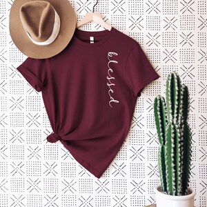 May include: A maroon t-shirt with the word "blessed" in white script. The shirt is hanging on a wooden hanger, next to a brown hat with a white band. A potted cactus sits to the right.