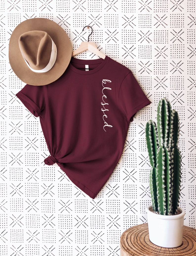 May include: A maroon t-shirt with the word "blessed" in white script. The shirt is hanging on a wooden hanger, next to a brown hat with a white band. A potted cactus sits to the right.