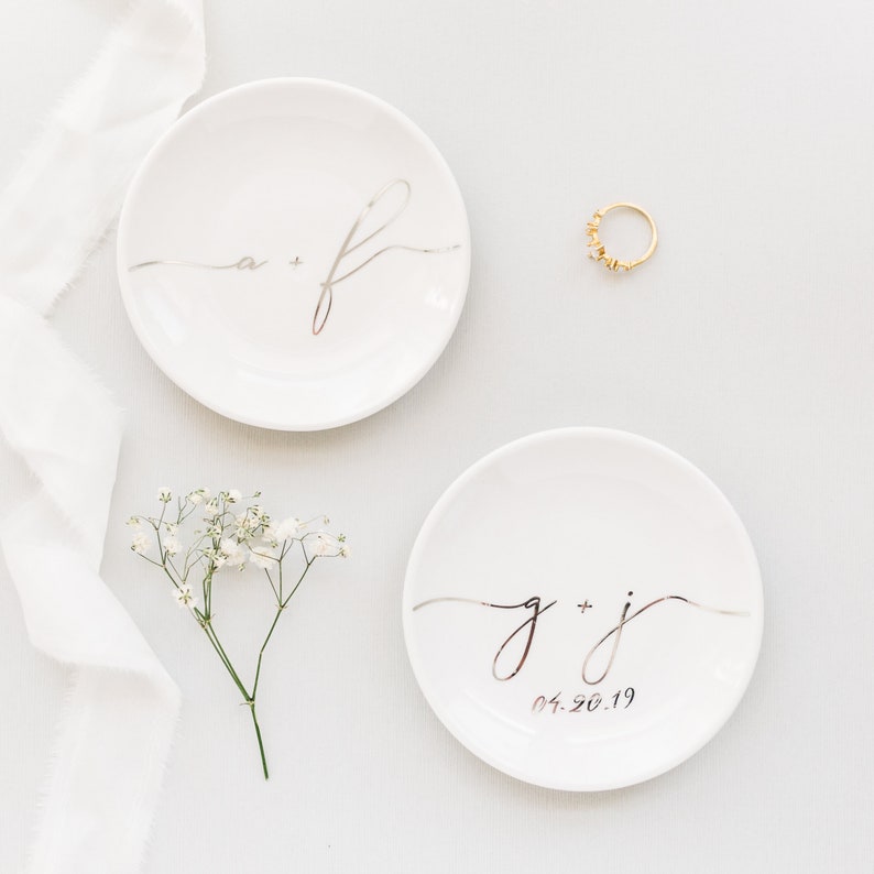 May include: Two white ceramic ring dishes with silver lettering. The top dish has the initials "a + f" and the bottom dish has the initials "g + j" and the date "04.26.19".