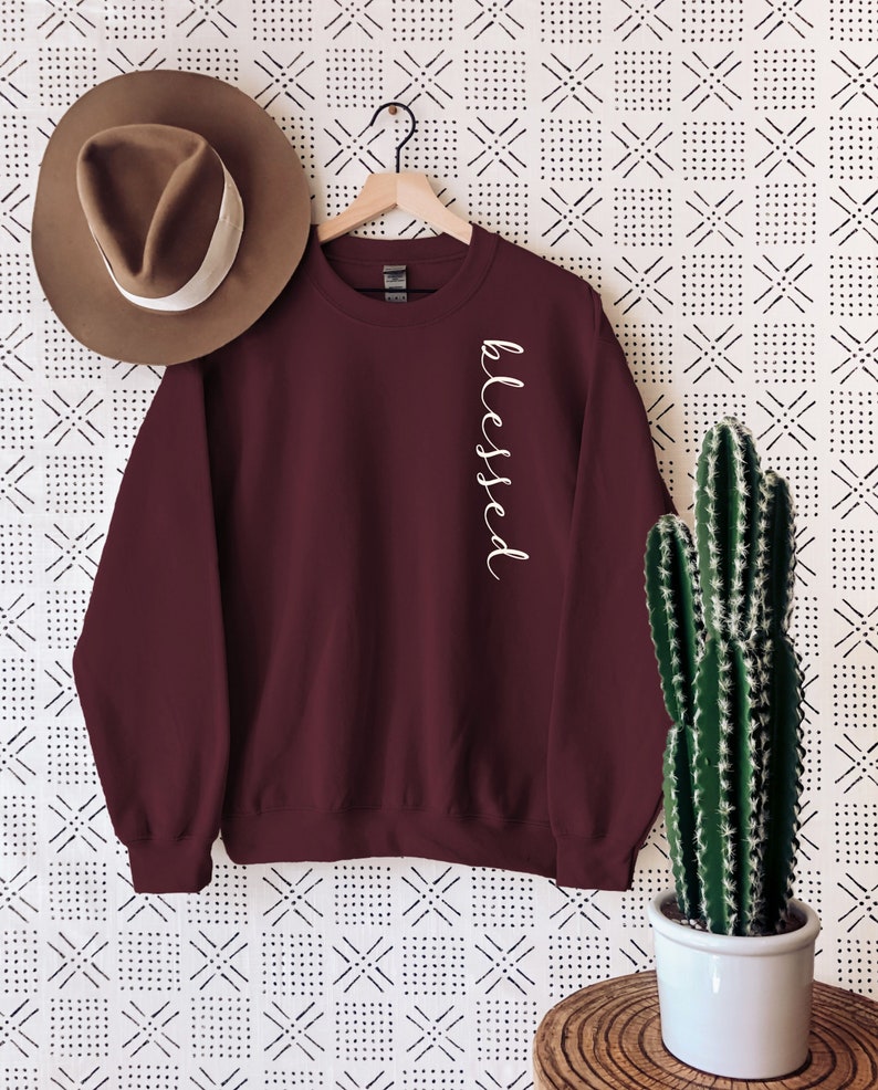 May include: A maroon sweatshirt with the word "blessed" in white script down the side. A brown hat with a white band hangs on the left. A cactus in a white pot sits on a wooden table to the right.