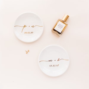 Peut inclure: Deux coupelles &agrave; anneaux en c&eacute;ramique blanche avec inscription dor&eacute;e. La premi&egrave;re coupelle indique "p + n 09.05.20" et la seconde "m + d 12.25.18". Un petit flacon de parfum avec une &eacute;tiquette dor&eacute;e indiquant "Gold Moon Perfume" est en arri&egrave;re-plan.
