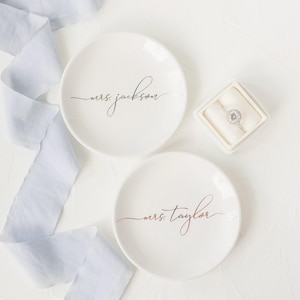 Puede incluir: Dos platos de cerámica blancos con texto personalizado. Uno tiene "mrs. jackson" en escritura negra, el otro "mrs. taylor" en escritura oro rosa. También hay un anillo de diamantes en una caja blanca.