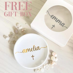 May include: A white ceramic ring dish with the name "amelia" in gold script and a gold cross. The dish is surrounded by small stones and a gift box with the name "emma" and a cross. Text in the image reads "FREE GIFT BOX".