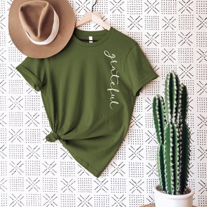 May include: Olive green t-shirt with the word "grateful" in white script. A brown fedora hat with a white band rests on the shirt. A cactus in a white pot is on the right. The background is a white and black geometric pattern.