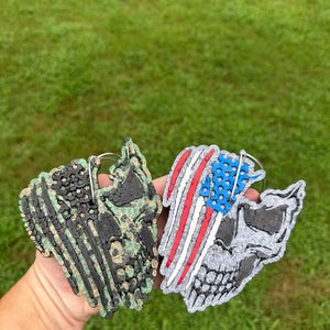 May include: Two skull-shaped air fresheners. One features a camouflage pattern, the other a design of the American flag. Both have silver metal loops for hanging. The background is green grass.