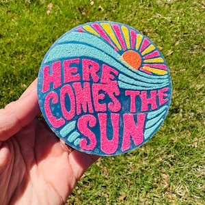May include: A round, blue, yellow, and pink coaster with the text "Here Comes The Sun" in pink letters. The sun is depicted in orange and yellow.