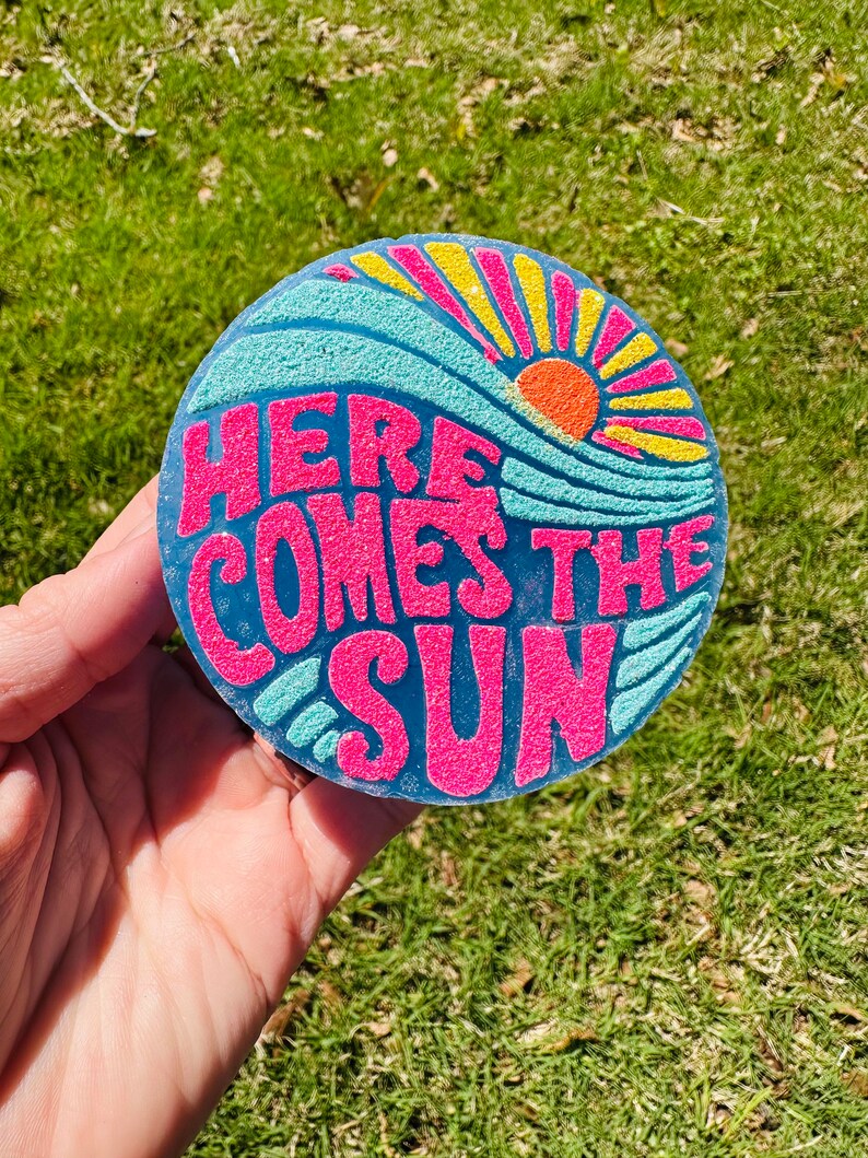 May include: A round, blue, yellow, and pink coaster with the text "Here Comes The Sun" in pink letters. The sun is depicted in orange and yellow.