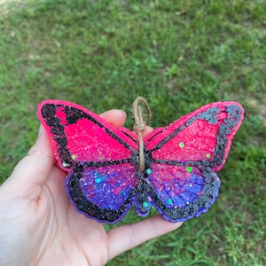 May include: A pink and purple glitter butterfly shaped wax melt with a black outline. The wax melt has a string attached for hanging.