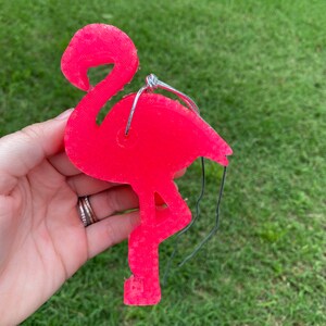 May include: A bright pink flamingo ornament with a wire hanger. The flamingo is made of a textured material and has a detailed design. The ornament is a vibrant shade of pink, perfect for adding a touch of tropical flair.