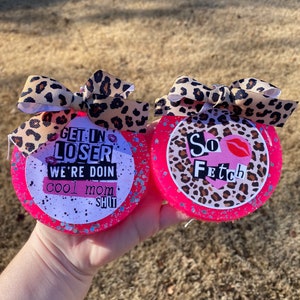 May include: Two pink round coasters with glitter and a leopard print design. One coaster says "Get in Loser We're Doin Cool Mom Shit" and the other says "So Fetch".